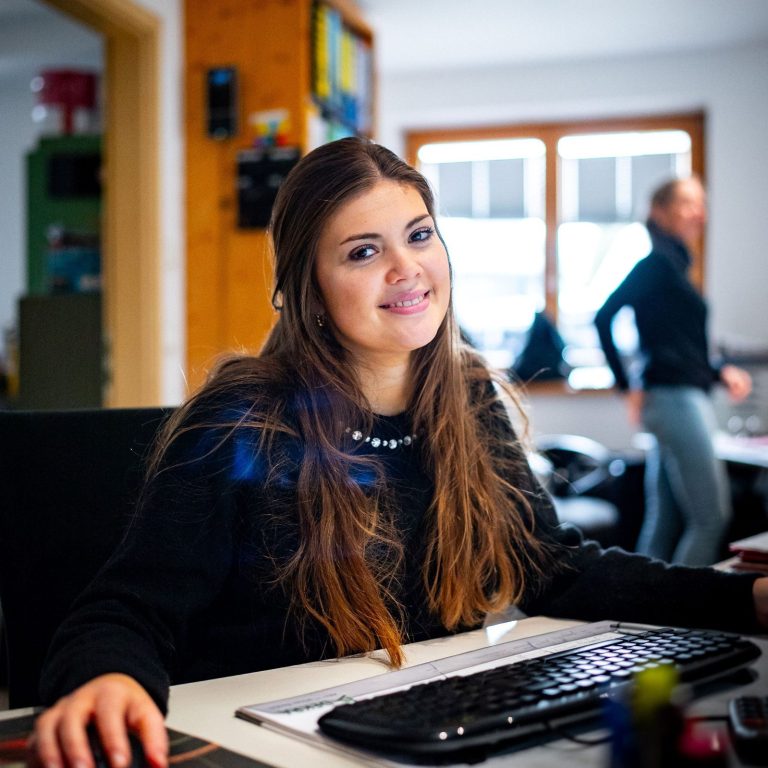 Junge Frau lächelt am Schreibtisch, im Hintergrund eine weitere Person bei der Arbeit.