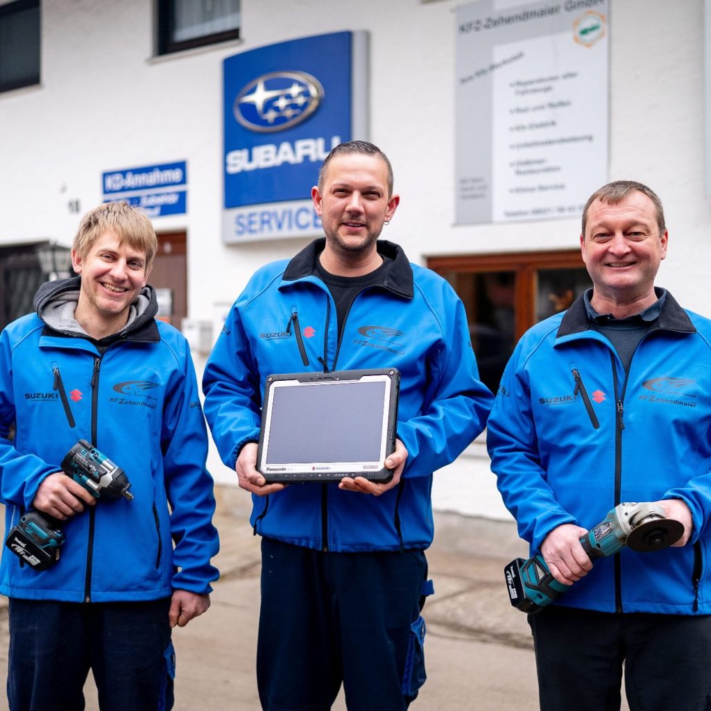 Drei KFZ-Meister in blauen Jacken halten Werkzeuge und ein Tablet vor der KFZ-Werkstatt.