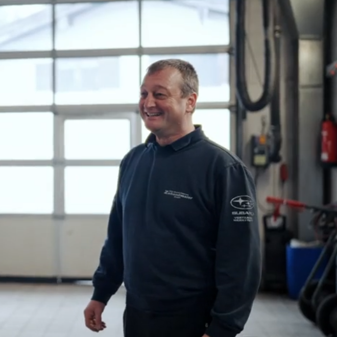 Lächelnder Chef in einem Werkstattumfeld, trägt einen dunkelblauen Pullover.