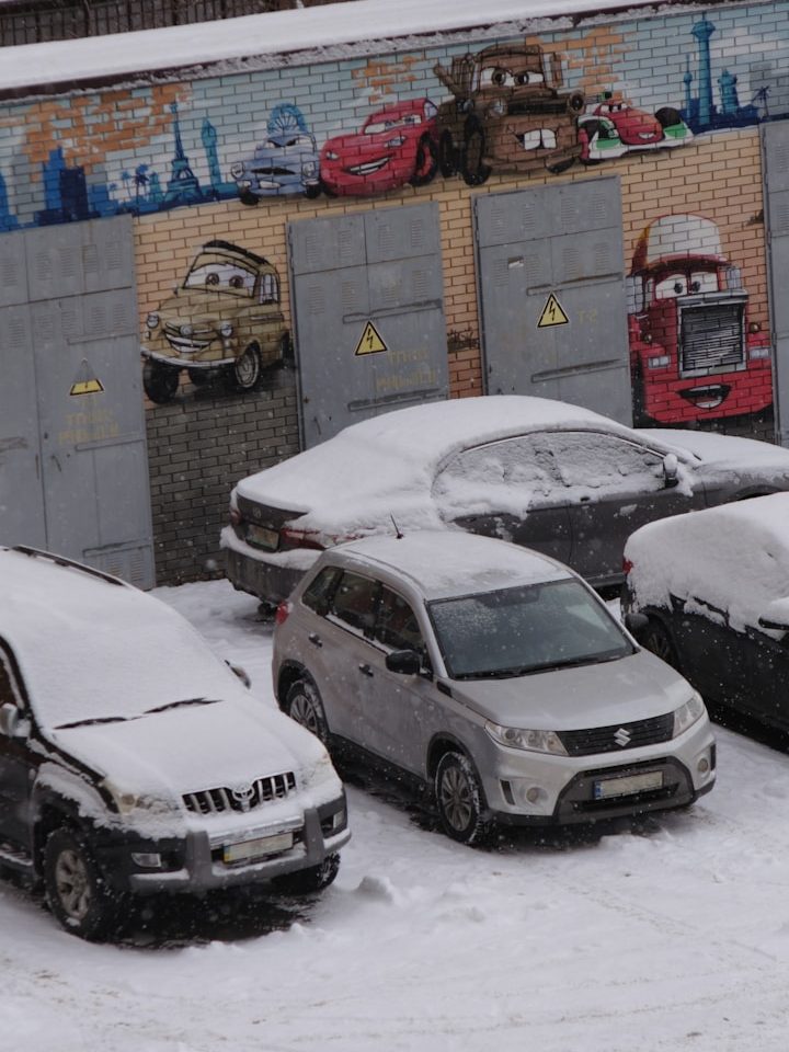 Verschneite Autos auf einem Parkplatz, mit einer Wandmalerei im Hintergrund. Das Auto mit Standheizung hat freie Scheiben.