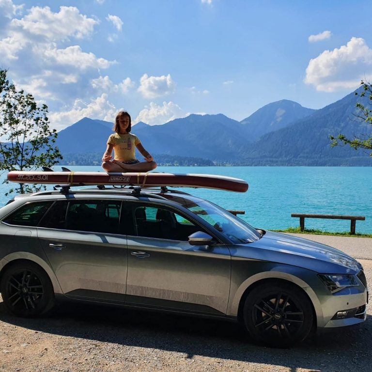 Mädchen sitzt im Lotussitz auf einem Stand-Up-Paddle-Board, das wiederum auf dem Autodach montiert ist. Im Hintergrund der Walchensee.