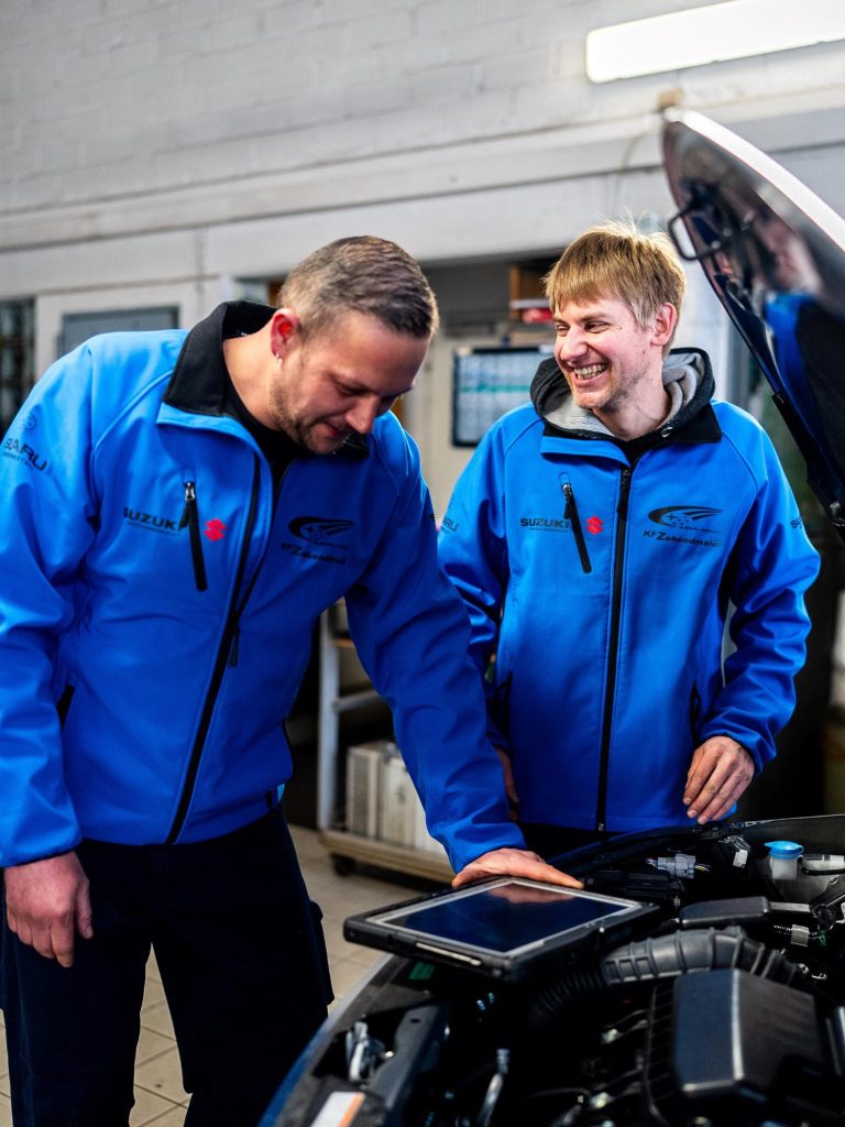 Zwei Mechanikermeister in blauen Jacken führen einen Systemcheck bei einem  Fahrzeug durch. Die Motorhaube ist geöffnet. 