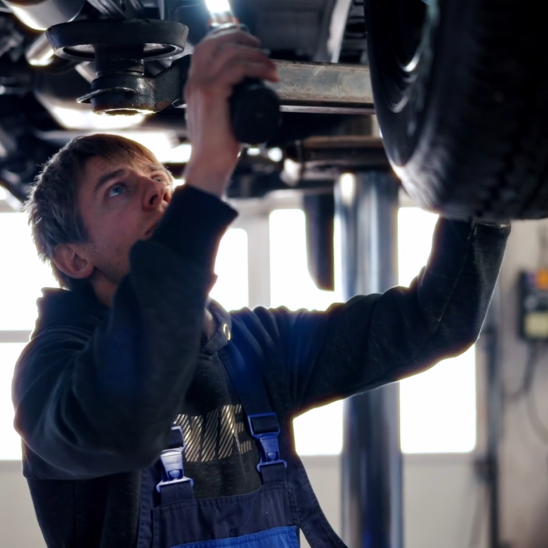 Junger Mechanikermeister arbeitet unter einem Auto mit einem Werkzeug.