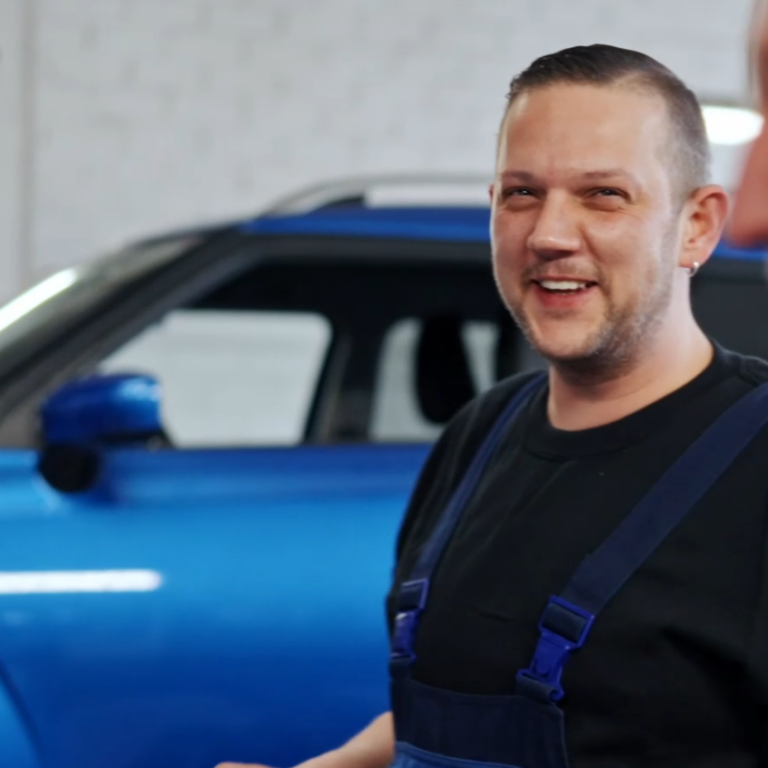 Der Werkstattleiter lächelt vor einem blauen Auto in einer Werkstatt.