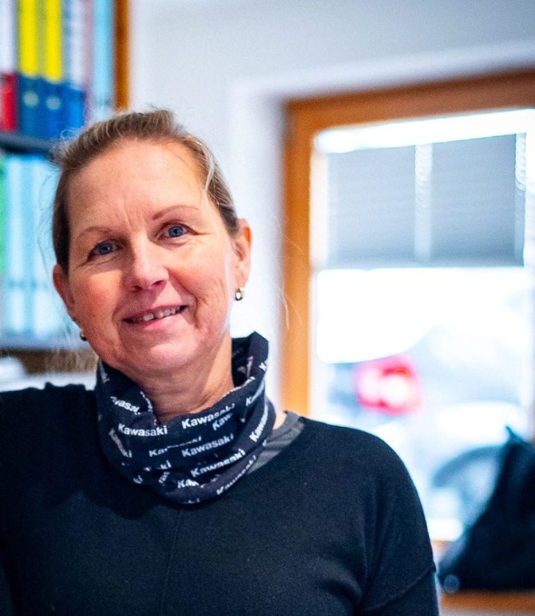 Frau mit lächelndem Gesicht, trägt einen Schal und steht in einem Büro.