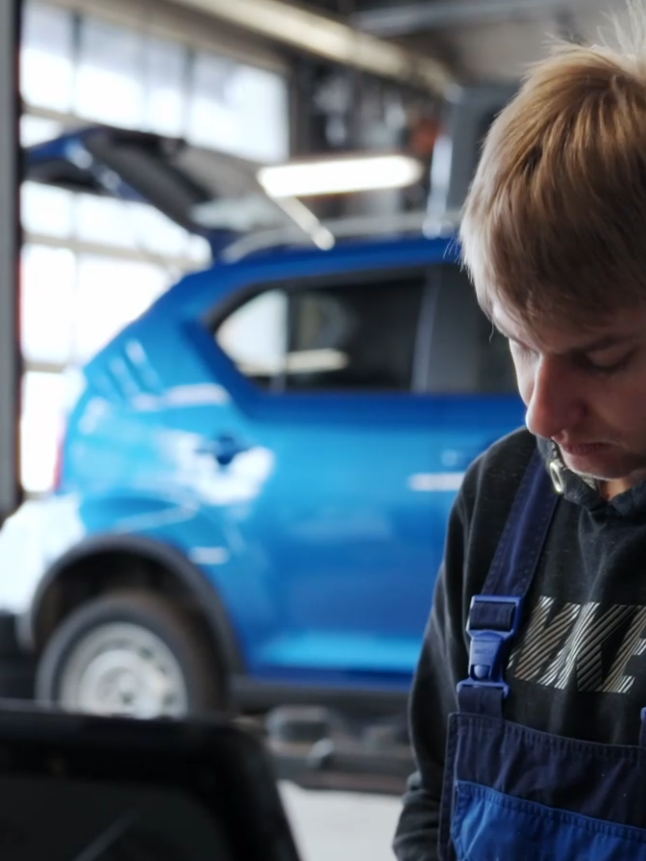Mechaniker arbeitet an einem Fahrzeug in einer Werkstatt mit blauem Auto im Hintergrund.