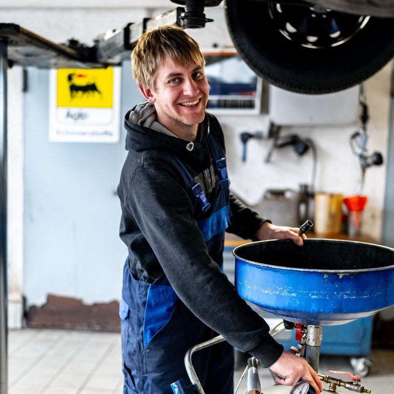 Junger KFZ-Meister in blauer Latzhose, der in der Werkstatt gerade einen Ölwechsel macht.